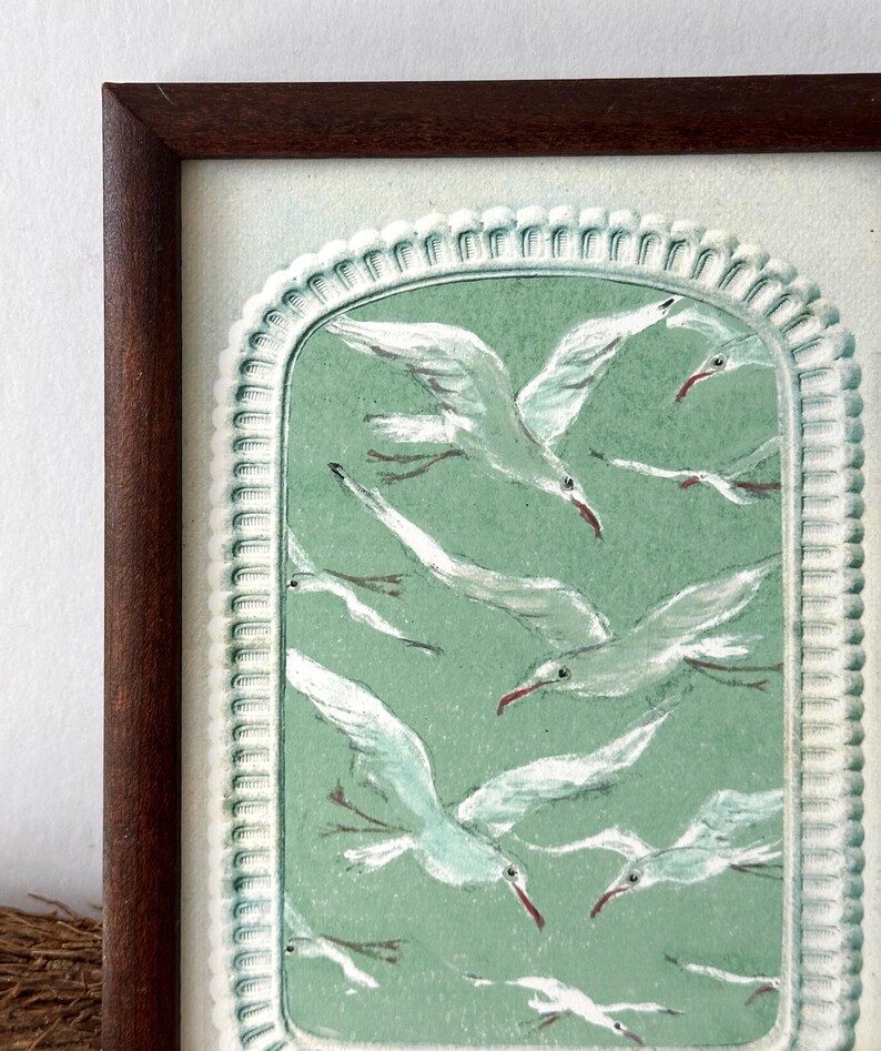 Puede incluir: Una obra de arte enmarcada que presenta una bandada de gaviotas blancas con picos rojos volando sobre un fondo verde claro. La obra est&aacute; encerrada en un borde blanco decorativo y un marco de madera marr&oacute;n oscuro, creando una est&eacute;tica vintage.