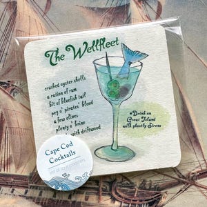 May include: A square coaster featuring a watercolor illustration of a cocktail glass with olives and a blue fish tail. Text reads The Wellfleet, a list of ingredients, and Drink on Great Island with ghostly Sirens. A round sticker says Cape Cod Cocktails.