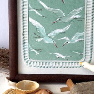 Puede incluir: Una obra de arte enmarcada que representa una bandada de gaviotas blancas con picos rojos sobre un fondo verde azulado apagado. La obra de arte est&aacute; en un marco de madera marr&oacute;n oscuro. Conchas marinas y una peque&ntilde;a figura de p&aacute;jaro de madera en primer plano.