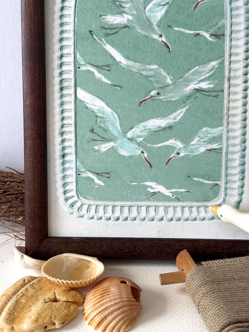 Puede incluir: Una obra de arte enmarcada que representa una bandada de gaviotas blancas con picos rojos sobre un fondo verde azulado apagado. La obra de arte est&aacute; en un marco de madera marr&oacute;n oscuro. Conchas marinas y una peque&ntilde;a figura de p&aacute;jaro de madera en primer plano.