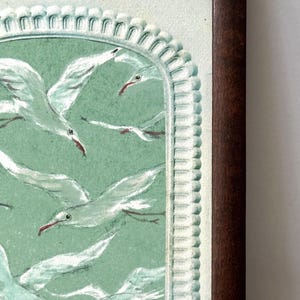 Puede incluir: Obra de arte enmarcada que presenta una bandada de gaviotas blancas con picos rojos sobre un fondo verde claro. La obra est&aacute; enmarcada en un marco de madera marr&oacute;n oscuro con un borde decorativo. Las aves est&aacute;n en vuelo, creando una sensaci&oacute;n de movimiento.