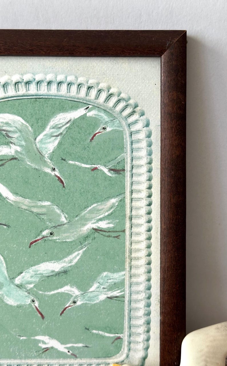 Puede incluir: Obra de arte enmarcada que presenta una bandada de gaviotas blancas con picos rojos sobre un fondo verde claro. La obra est&aacute; enmarcada en un marco de madera marr&oacute;n oscuro con un borde decorativo. Las aves est&aacute;n en vuelo, creando una sensaci&oacute;n de movimiento.