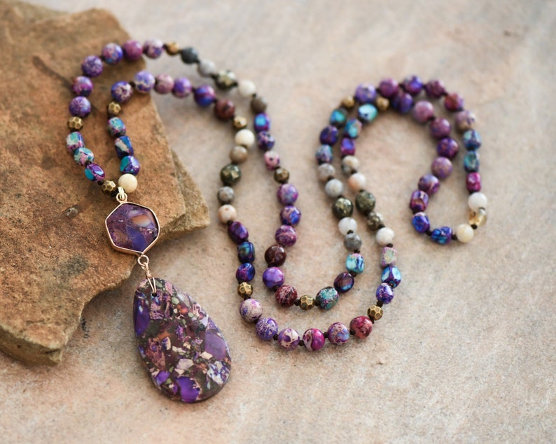 Peut inclure: Un long collier de perles violettes, bleues et dor&eacute;es avec un grand pendentif violet et dor&eacute;. Le collier est fait de pierres naturelles et est parfait pour &ecirc;tre superpos&eacute;.