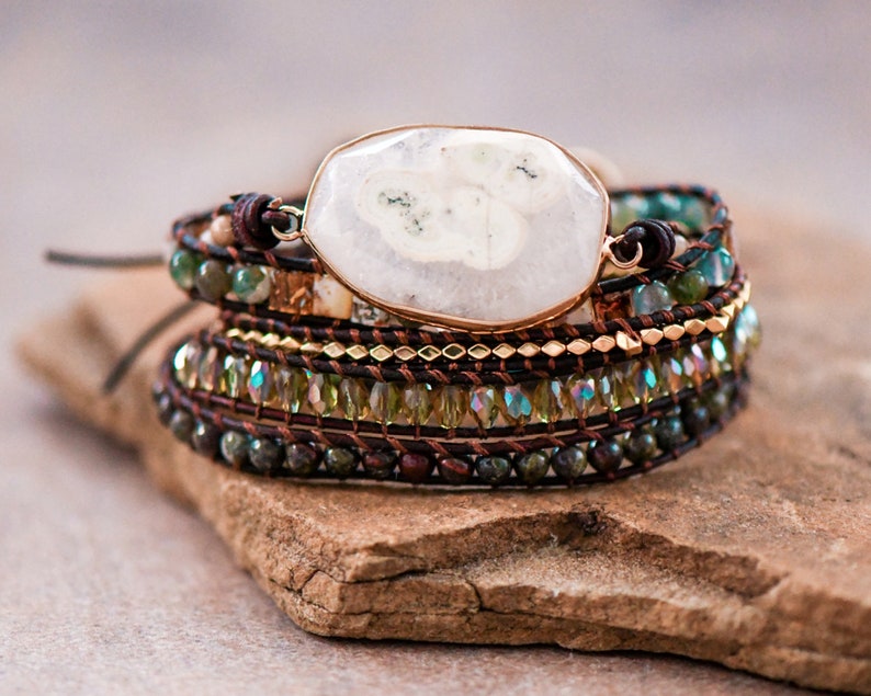 May include: A multi-layered bracelet with a large white agate gemstone, brown leather cord, and green and gold beads.