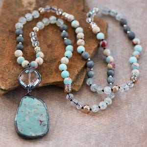 May include: A handmade necklace with a large, irregular turquoise pendant in a dark metal frame. The necklace includes a clear crystal pendant and strands of grey, blue, and clear beads, with gold-coloured beads. The necklace is displayed on a textured, brown surface.