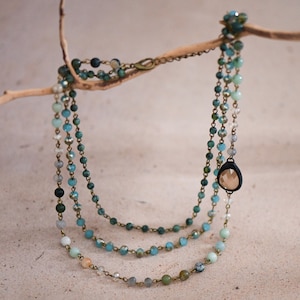 Puede incluir: Un collar de varias capas con cuentas turquesa, verdes y blancas, y un colgante en forma de lágrima con un cristal transparente.