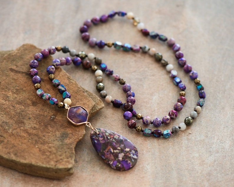 Peut inclure: Un collier multi-rangs avec des perles violettes, bleues et blanches. Le collier a un grand pendentif en pierre violette et blanche.