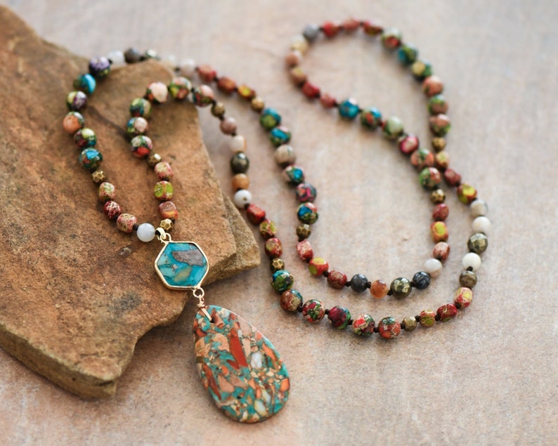 May include: A long necklace made of colorful beads with a turquoise and brown stone pendant. The necklace is on a brown textured surface.