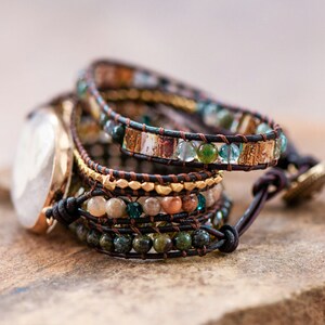 May include: Multi-layered bracelet with brown leather straps and colorful beads, including green, blue, and gold. The bracelet has a decorative clasp.