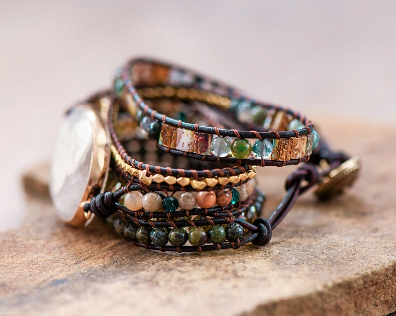 May include: Multi-layered bracelet with brown leather straps and colorful beads, including green, blue, and gold. The bracelet has a decorative clasp.