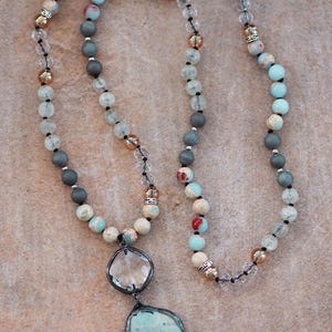 May include: A long necklace with a large, irregular turquoise pendant in a silver-tone frame. The necklace features beads in blue, grey, cream, and clear, with metallic accents. The beads are strung on a black cord. The necklace is a piece of jewellery.