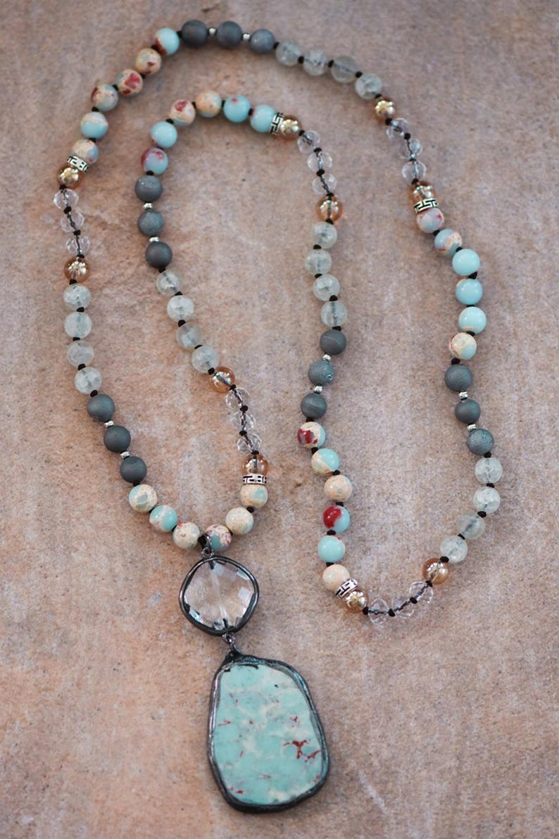 May include: A long necklace with a large, irregular turquoise pendant in a silver-tone frame. The necklace features beads in blue, grey, cream, and clear, with metallic accents. The beads are strung on a black cord. The necklace is a piece of jewellery.