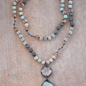 May include: A layered necklace with a large, irregular turquoise pendant and a clear, faceted stone pendant. The necklace has multiple strands of beads in shades of blue, grey, and cream, with clear crystal accents. The beads are strung on a black cord.