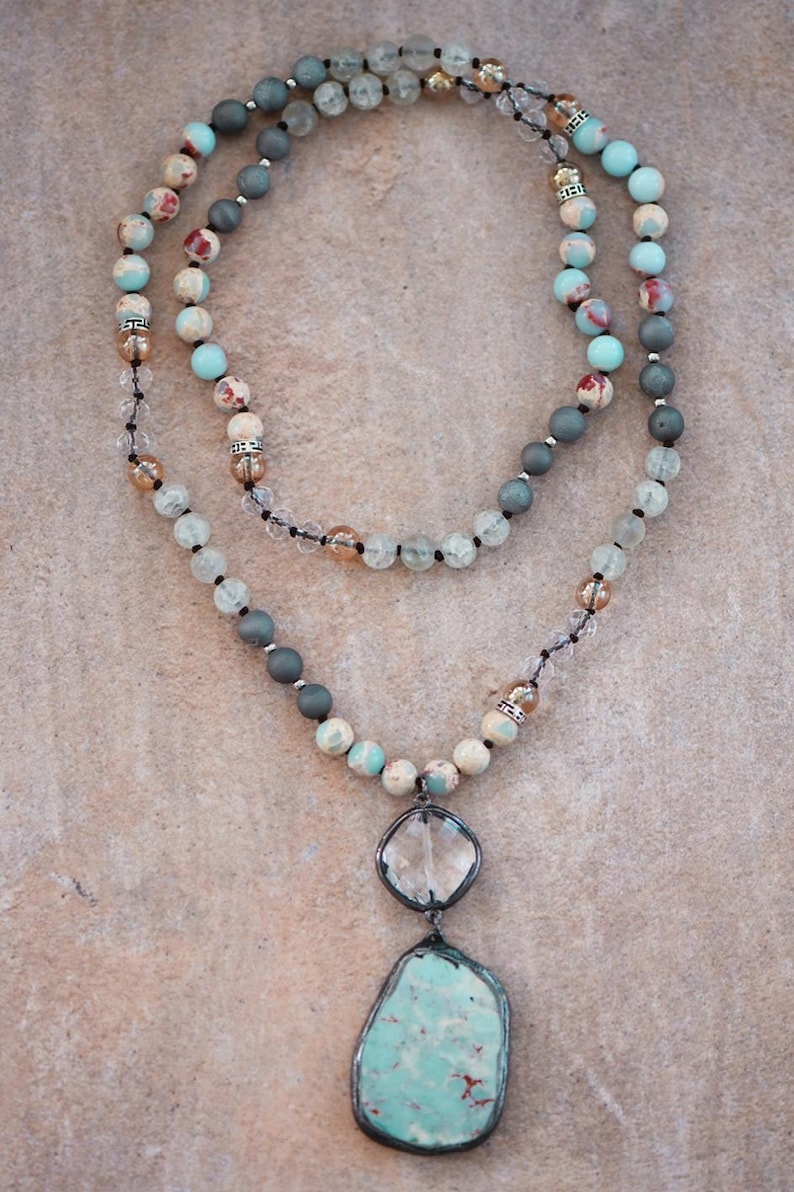 May include: A layered necklace with a large, irregular turquoise pendant and a clear, faceted stone pendant. The necklace has multiple strands of beads in shades of blue, grey, and cream, with clear crystal accents. The beads are strung on a black cord.