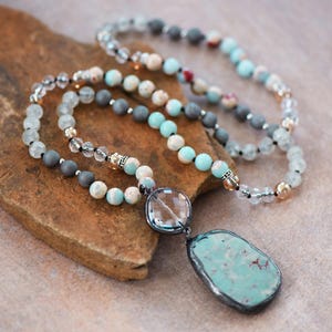 May include: A layered necklace with beads in grey, cream, and turquoise. The necklace includes clear crystal beads and a large, irregular turquoise pendant with a silver-coloured border. The necklace is displayed on a brown, textured surface.