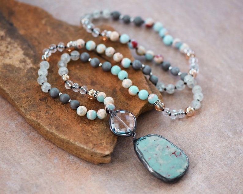 May include: A layered necklace with beads in grey, cream, and turquoise. The necklace includes clear crystal beads and a large, irregular turquoise pendant with a silver-coloured border. The necklace is displayed on a brown, textured surface.
