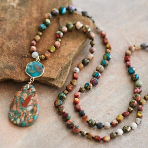 May include: A multi-strand necklace with a large, teardrop-shaped pendant. The necklace is made of small, colorful beads and features a mix of gold, brown, red, blue, and green tones. The pendant is a mix of brown, red, and blue.