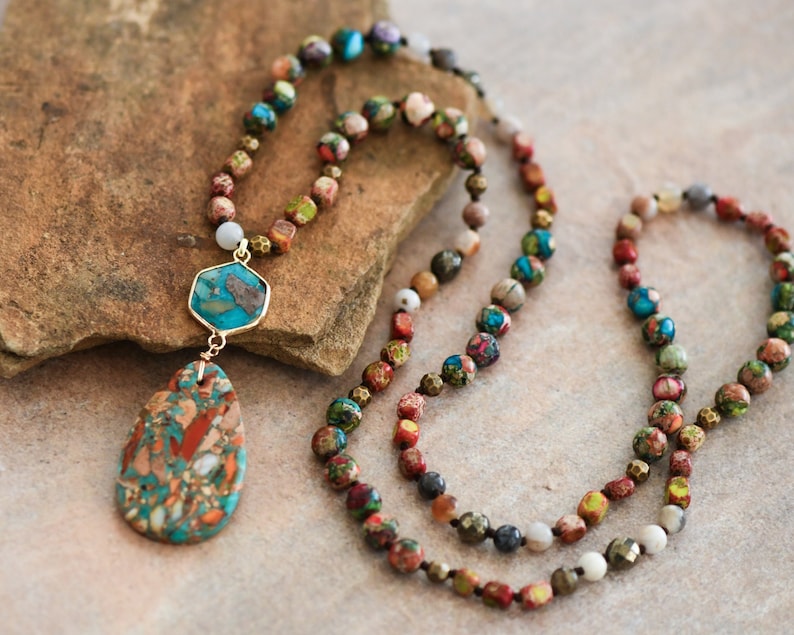 May include: A multi-strand necklace with a large, teardrop-shaped pendant. The necklace is made of small, colorful beads and features a mix of gold, brown, red, blue, and green tones. The pendant is a mix of brown, red, and blue.