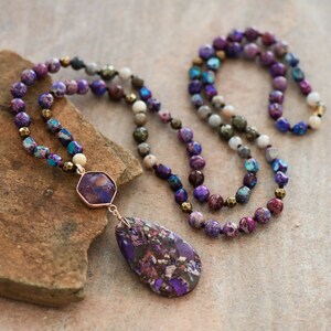 Peut inclure: Un long collier de perles violettes, bleues et brunes avec un grand pendentif violet et dor&eacute;. Le collier est sur une roche brune.
