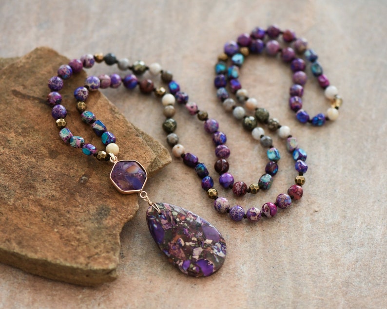 Peut inclure: Un long collier de perles violettes, bleues et brunes avec un grand pendentif violet et dor&eacute;. Le collier est sur une roche brune.
