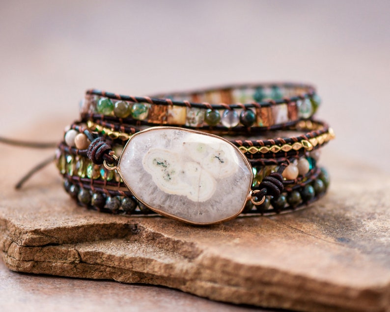 May include: A multi-layered wrap bracelet with a large white agate stone pendant. The bracelet features brown leather cord, gold beads, and green and brown gemstone beads.