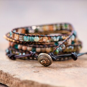 May include: A multi-layered wrap bracelet with brown leather cord and various colored beads, including green, brown, and white. The bracelet has a decorative metal clasp with a floral design.