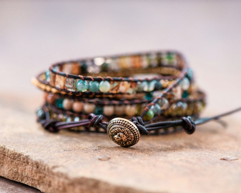 May include: A multi-layered wrap bracelet with brown leather cord and various colored beads, including green, brown, and white. The bracelet has a decorative metal clasp with a floral design.