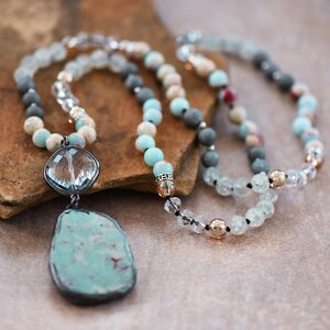 May include: Two beaded necklaces are displayed. One has a large, turquoise pendant with a silver frame. The other features beads in blue, grey, and clear, with gold-coloured accents. The necklaces are displayed on a brown surface.