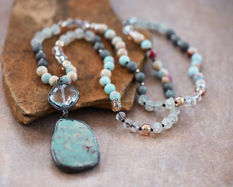 May include: Two beaded necklaces are displayed. One has a large, turquoise pendant with a silver frame. The other features beads in blue, grey, and clear, with gold-coloured accents. The necklaces are displayed on a brown surface.