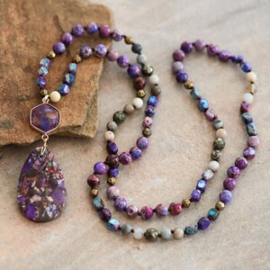 Peut inclure: Un long collier de perles violettes et grises avec un pendentif en pierre pr&eacute;cieuse violette. Le pendentif est en forme de larme avec une plus petite pierre pr&eacute;cieuse hexagonale au-dessus.