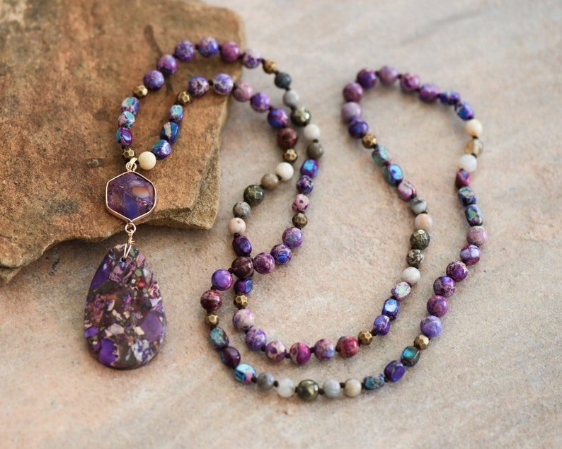 Peut inclure: Un long collier de perles violettes et grises avec un pendentif en pierre pr&eacute;cieuse violette. Le pendentif est en forme de larme avec une plus petite pierre pr&eacute;cieuse hexagonale au-dessus.