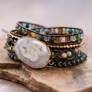 May include: A multi-layered bracelet with a large, white agate stone charm. The bracelet is made with brown leather cord, gold beads, and green and brown stones.