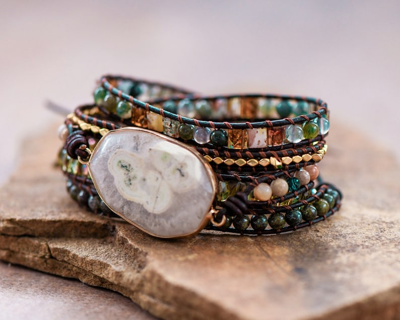 May include: A multi-layered bracelet with a large, white agate stone charm. The bracelet is made with brown leather cord, gold beads, and green and brown stones.