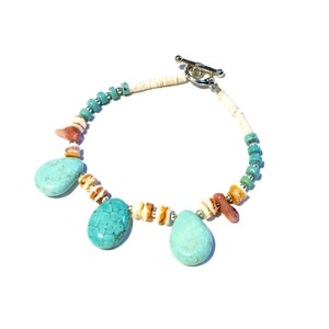 May include: A turquoise and shell beaded bracelet with a silver toggle clasp. The bracelet features three teardrop-shaped turquoise stones and is accented with small turquoise beads and shell fragments. The design is a mix of natural elements and a touch of elegance.