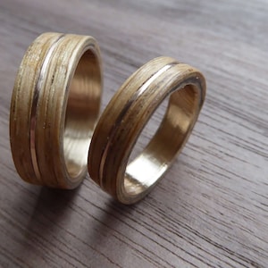 Puede incluir: Dos anillos de madera con finas bandas doradas. Los anillos están hechos de madera marrón claro con un patrón de veta natural.