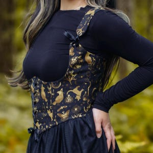 May include: A black long-sleeved top and a black skirt are worn with a decorative corset. The corset is black with a gold pattern of moons, stars, and animals. The outfit is worn outdoors in front of a blurred forest background.