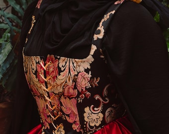 May include: A black corset with gold and red floral embroidery, laced with cream-colored ribbon, worn over a red satin skirt. The corset has a square neckline and is part of a historical costume.