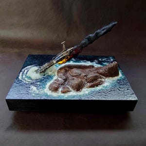 Puede incluir: Un diorama que muestra una nave espacial en miniatura estrellándose en un océano azul oscuro junto a una isla rocosa marrón. La nave está inclinada hacia abajo, emitiendo una estela de humo oscuro y llamas naranjas al impactar el agua.
