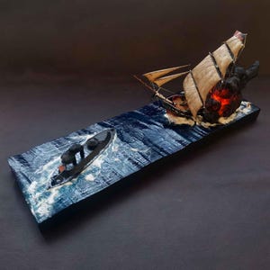 Può includere: Un modello di una nave in fiamme attaccata da un sottomarino nero più piccolo. La nave sta affondando e il sottomarino si sta dirigendo verso di essa. La scena è ambientata su una base di legno con un mare dipinto di blu e bianco.