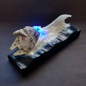 Könnte beinhalten: Ein Modell des Raumschiffs Millennium Falcon aus Star Wars, teilweise in einer Welle aus weißem Harz versunken. Das Modell ist mit blauen LED-Lichtern beleuchtet und steht auf einem schwarzen Holzsockel.