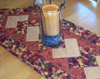 May include: A fall-themed table runner with a patchwork design featuring a brown, orange, and yellow leaf print, a tan gingham print, and a red floral print. The runner is accented with a decorative glass candle holder filled with blue beads.