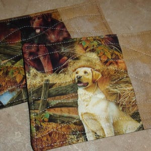 May include: Two quilted fabric coasters with a hunting theme. One coaster features a brown and black dog, while the other features a yellow dog in a field of tall grass.