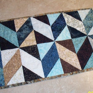 May include: A table runner with a geometric pattern in shades of blue, brown, and beige. The runner is made of fabric and has a brown border.