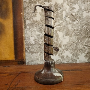 Könnte beinhalten: Ein antiker silberfarbener Kerzenhalter mit einem Spiraldesign. Der Sockel ist rund und dunkel, mit weißer Farbe. Der Kerzenhalter hat einen Haken oben und eine kleine Schlaufe an der Seite. Der Hintergrund zeigt ein Gemälde und eine Tapete mit Baummuster.