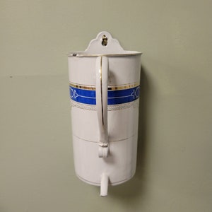 Può includere: Brocca per acqua da parete in smalto bianco con una fascia blu e dorata e un manico decorativo.