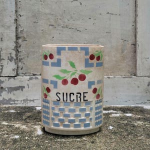 May include: A rectangular ceramic sugar container with a cream-colored base. The container features a blue and white geometric pattern, with red cherries and the word "SUCRE" in black lettering. The container is likely vintage.
