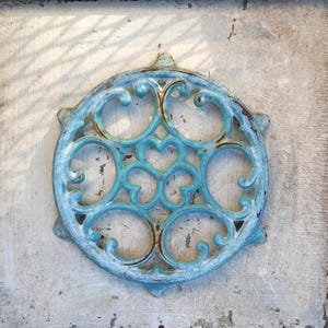 May include: A circular, turquoise cast iron decorative element with an intricate, openwork design. The piece, featuring heart and swirl patterns, is mounted on a weathered, white wooden surface. The metal has a distressed, aged appearance.