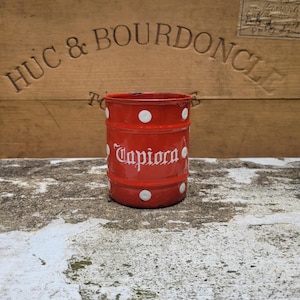 以下が含まれることがあります： 白い水玉模様と白い筆記体で「Tapioca」の文字が入った、ヴィンテージ風の赤い金属製容器。円筒形の容器は素朴で古びた外観をしており、アンティークまたはレトロなキッチン用品であると思われます。