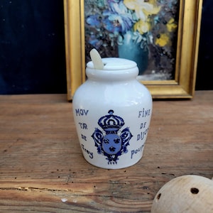 May include: A white ceramic jar with a lid and a small spoon. The jar has a blue and white crest design and text in French. The jar is sitting on a wooden surface.