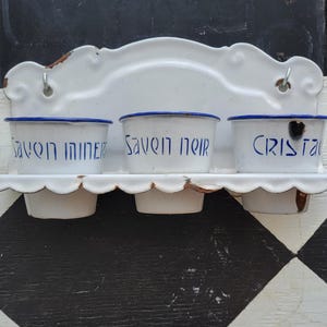 May include: A white enamel wall-mounted organizer with three small, round containers. Each container has a blue rim and blue text. The text on the containers reads "SAVON MINER", "SAVON NOIR", and "CRISTAL". The organizer has a scalloped shelf and decorative top.